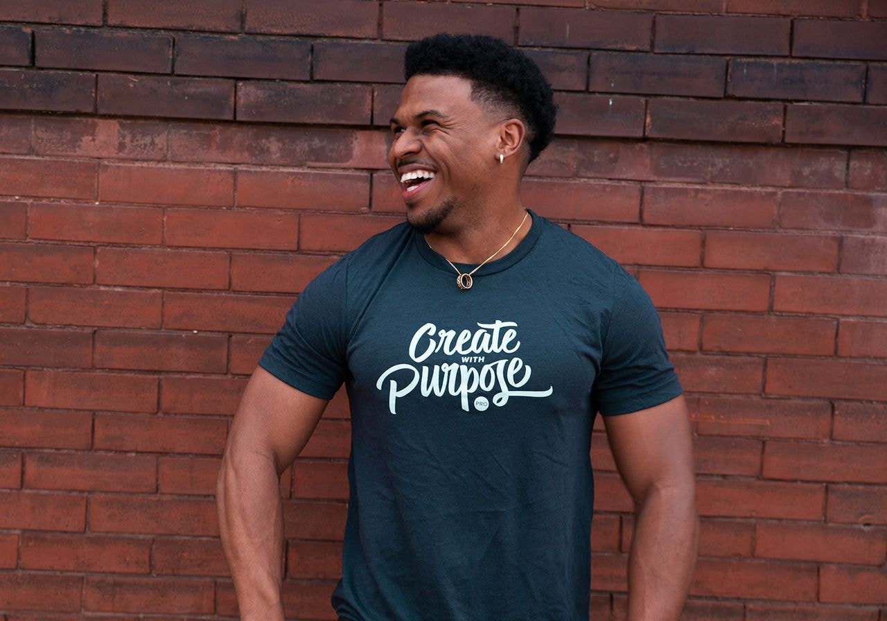 A smiling man in a blue “Create with purpose” t-shirt on a red brick background.